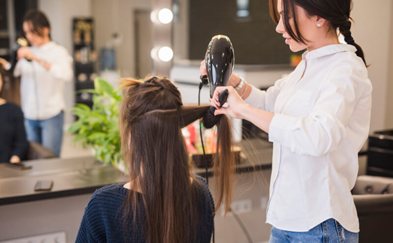 Cabeleireira trabalhando, segurando e secando o cabelo de cliente que esta sentada de costas